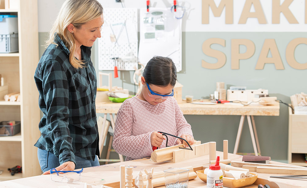 Lehrerin schaut Schülerin zu, wie sie mit der Handsäge Holz sägt